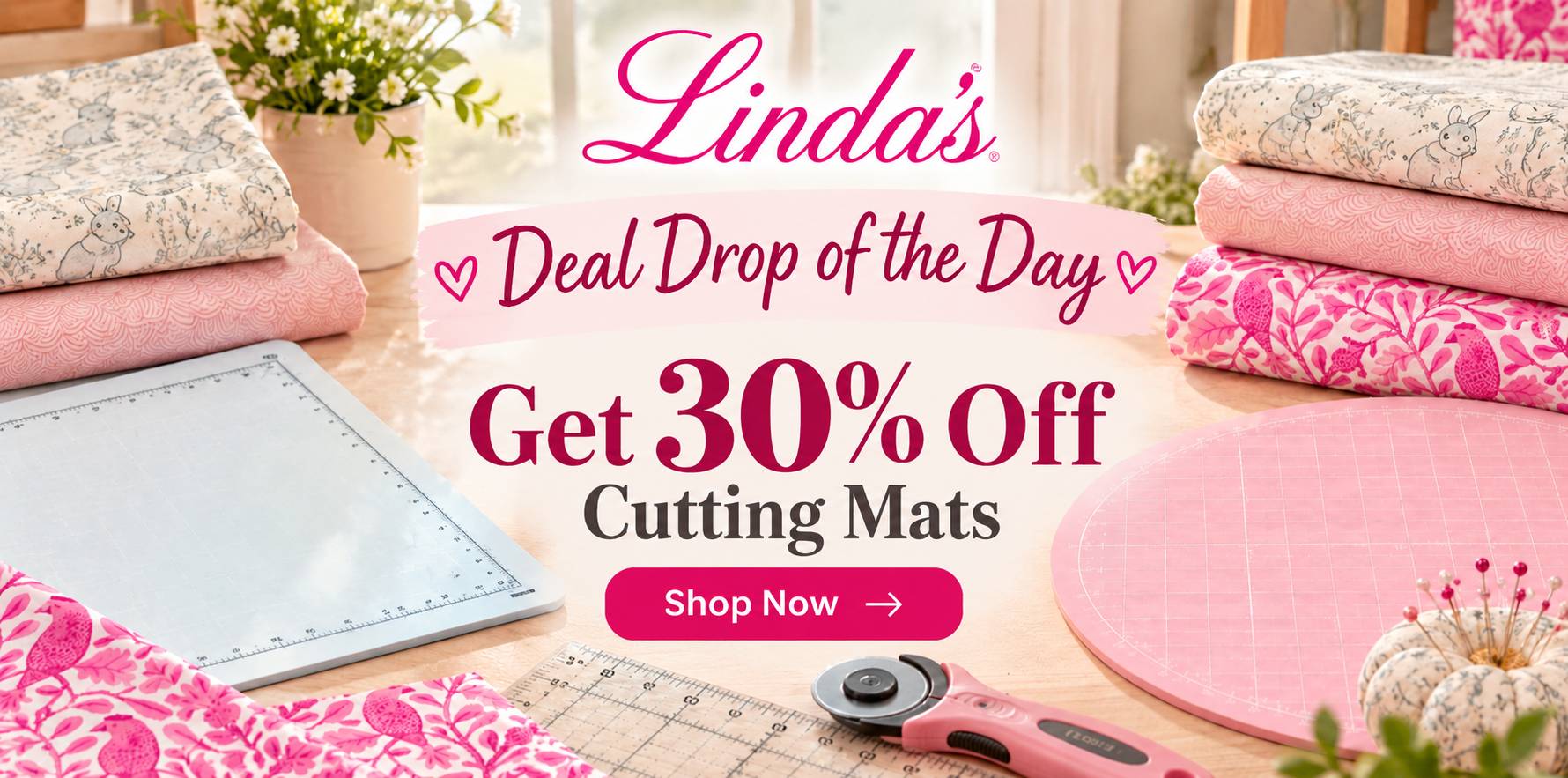 Wide, bright overhead-angled craft-table scene with folded cream and pink patterned fabrics, a gray rectangular cutting mat at left, a round pink grid...