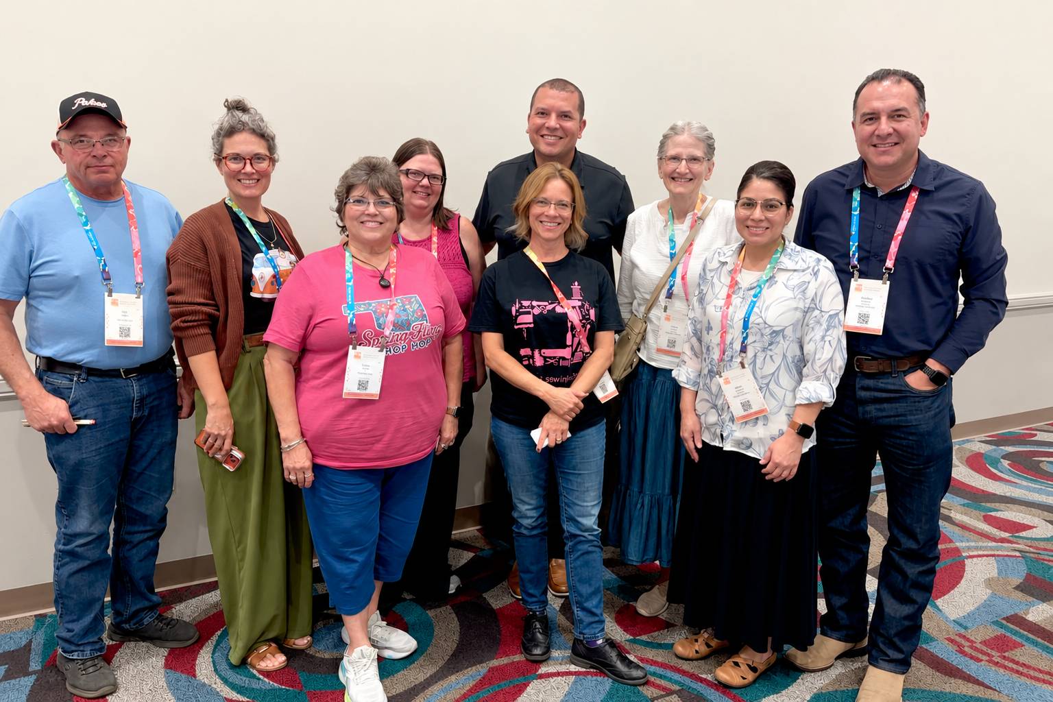 A wide, eye-level group portrait shows nine adults standing in two close rows against a plain cream wall, wearing casual shirts, jeans, skirts, confer...
