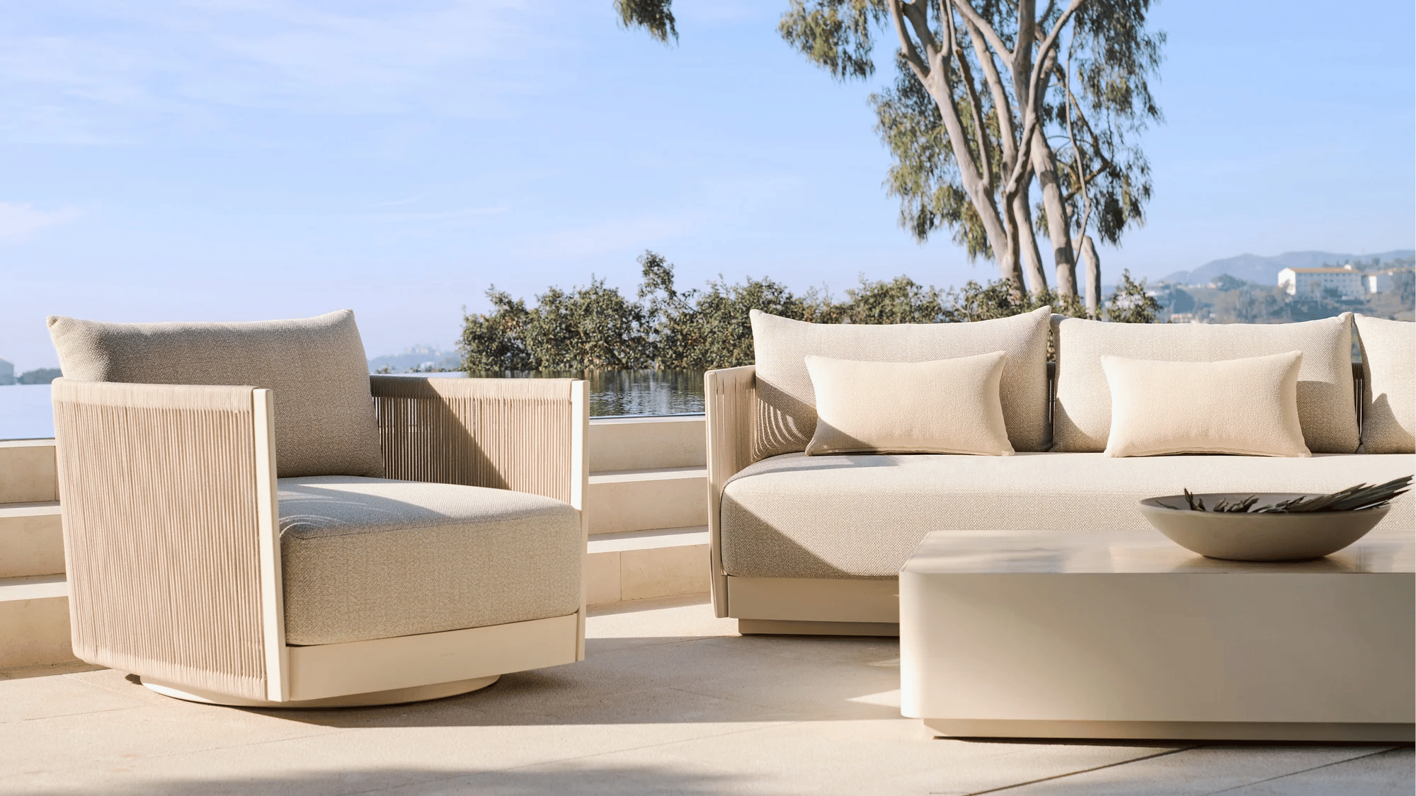 A medium shot captures a modern outdoor living space with a light-colored armchair and a sofa featuring beige cushions and pillows, positioned on a st...