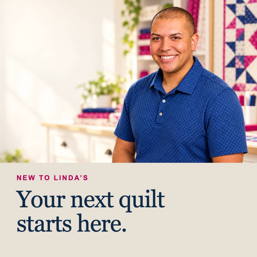 A medium-shot portrait shows a smiling person with closely cropped hair wearing a patterned blue collared shirt, seated before a bright sewing and qui...